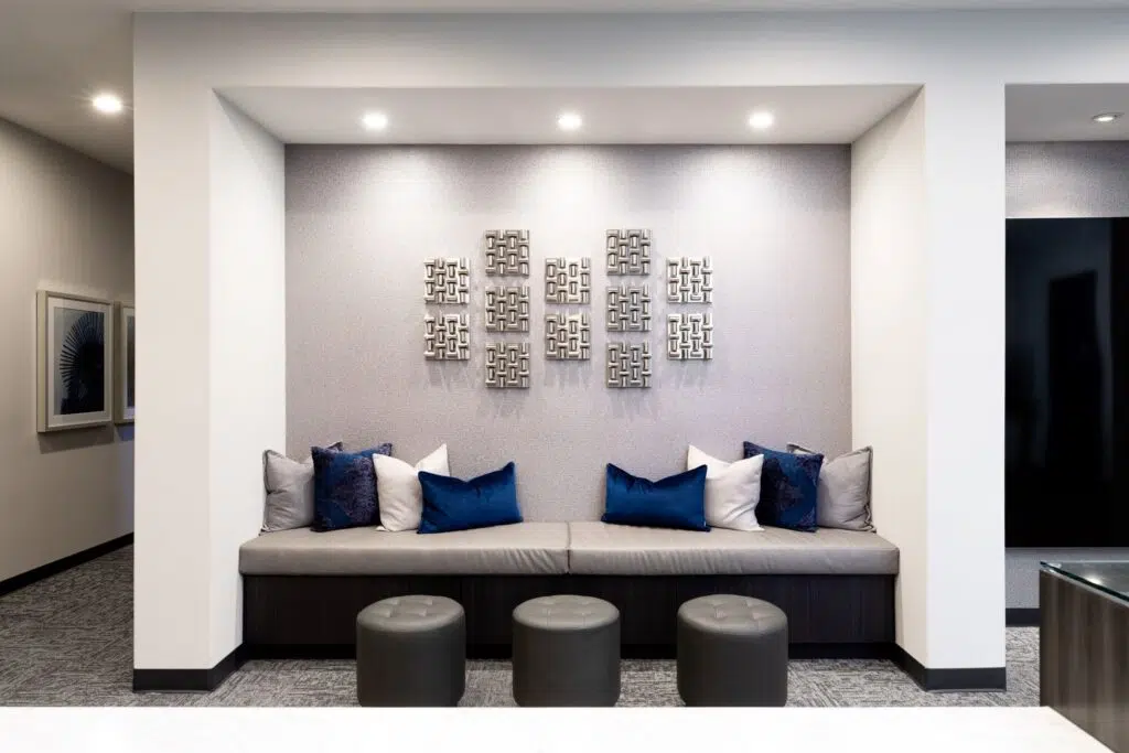 Seating area with a cubby-style bench featuring grey cushions and an array of grey and blue pillows on each side, three circular footrest chairs in front, silver square art pieces on the wall, and bright lighting.