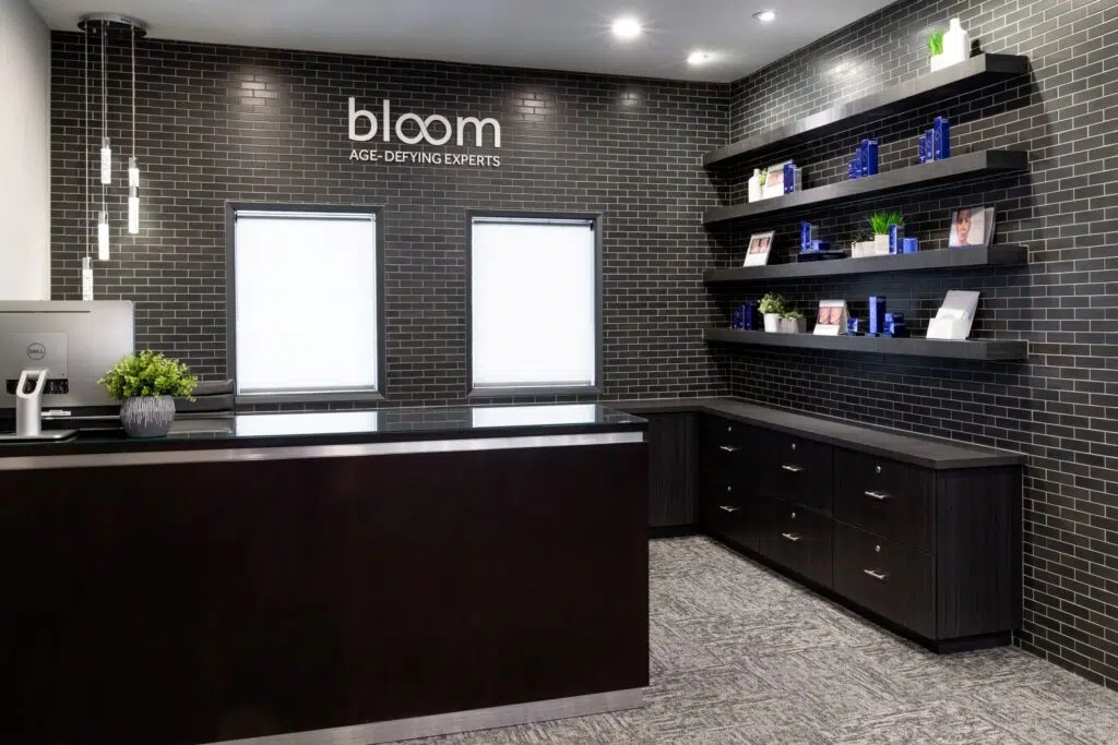 Bloom check-in and out desk at the Temecula location, with shelves on the right, Bloom logo above two windows, cascading ceiling lighting, a computer with a faux plant on the glass counter desk.