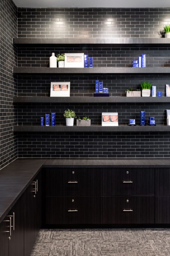 Four shelves on the wall in the Bloom section, featuring ZO products and faux plants, with dark grey rectangular tile on the wall and carpet flooring below, along with counters featuring drawers with silver handles.