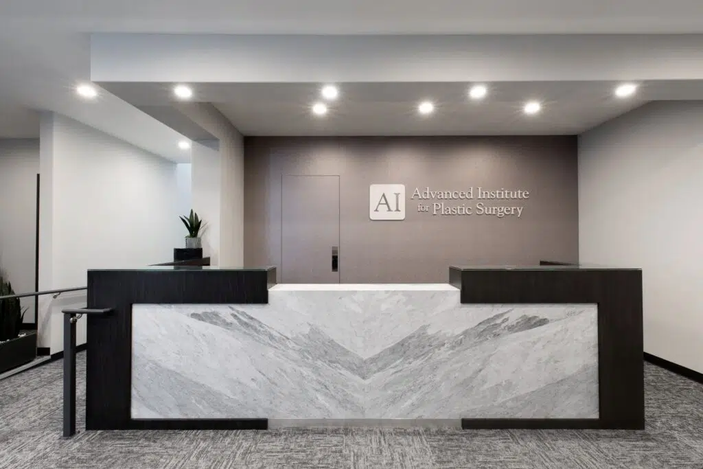 Checkout desk area at the Advanced Institute for Plastic Surgery, featuring a marble front counter, modern ceiling lighting, the logo on the back wall, and a faux plant on the counter with railings leading to the front lobby.