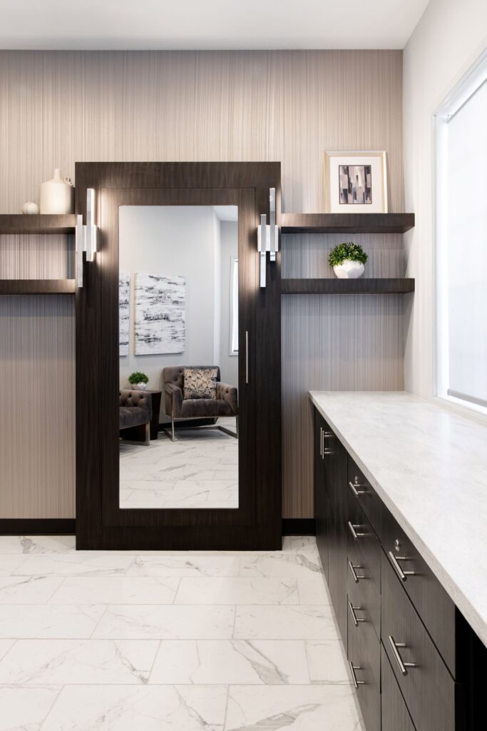Consultation room at the Advanced Institute for Plastic Surgery, featuring full-length mirrors, sleek grey design, modern decor, clean tile floors, and a marble countertop with drawers underneath with silver modern handles.