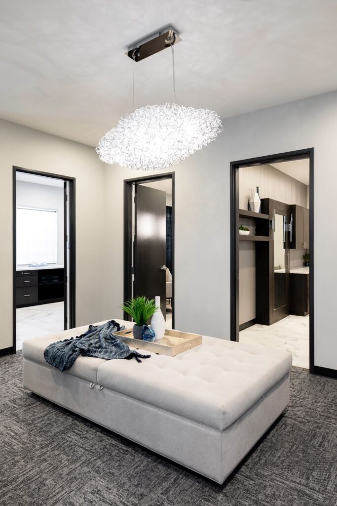 Elegant hallway leading to the consultation rooms at the Advanced Institute for Plastic Surgery, featuring a cloud-like light fixture, pristine consultation rooms, a flat cushion seating area with a gold tray and modern vases, one containing a faux plant, and a light blue throw blanket.